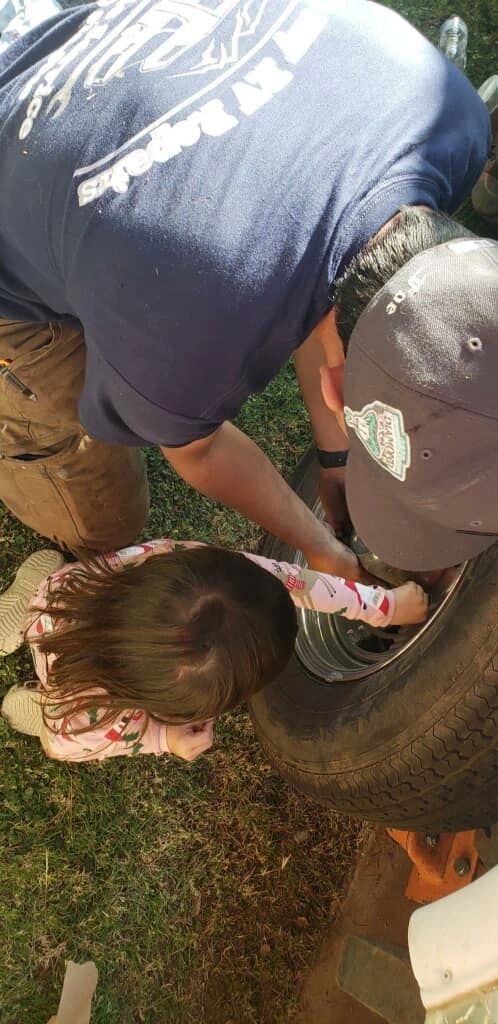 Technician in a Help My RV Repairs shirt working at an RV wheel hub on grass, with the wheel off and a jack under the axle.